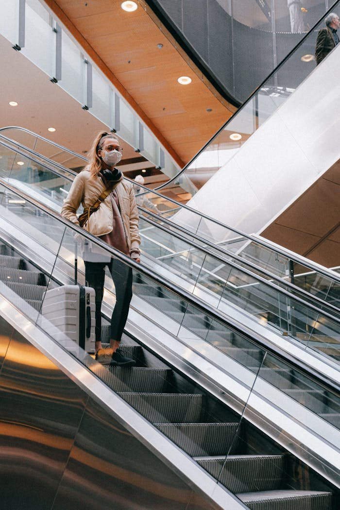 Woman with luggage wearing face mask on escalator in airport during pandemic.